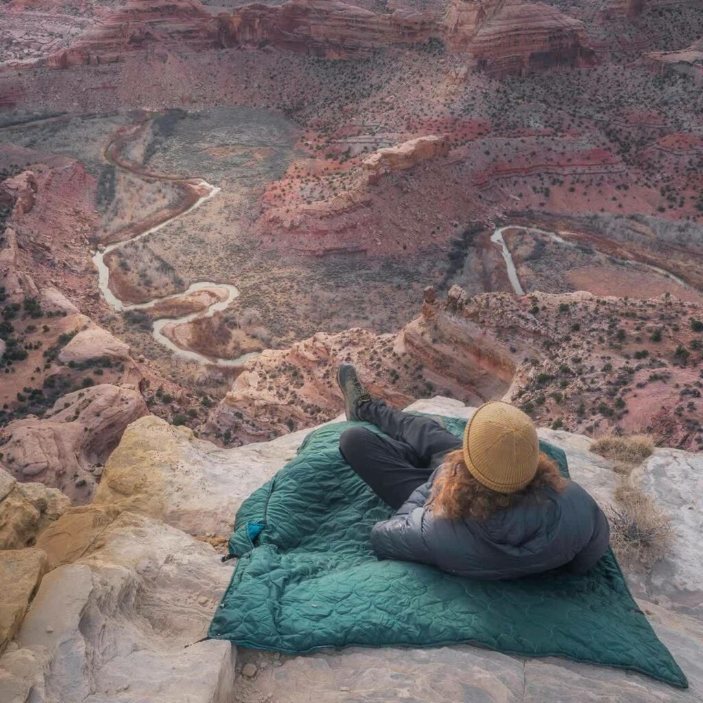 A person with curly red hair is lying down on a green blanket on a mountainside overlooking a dramatic rocky red valley.