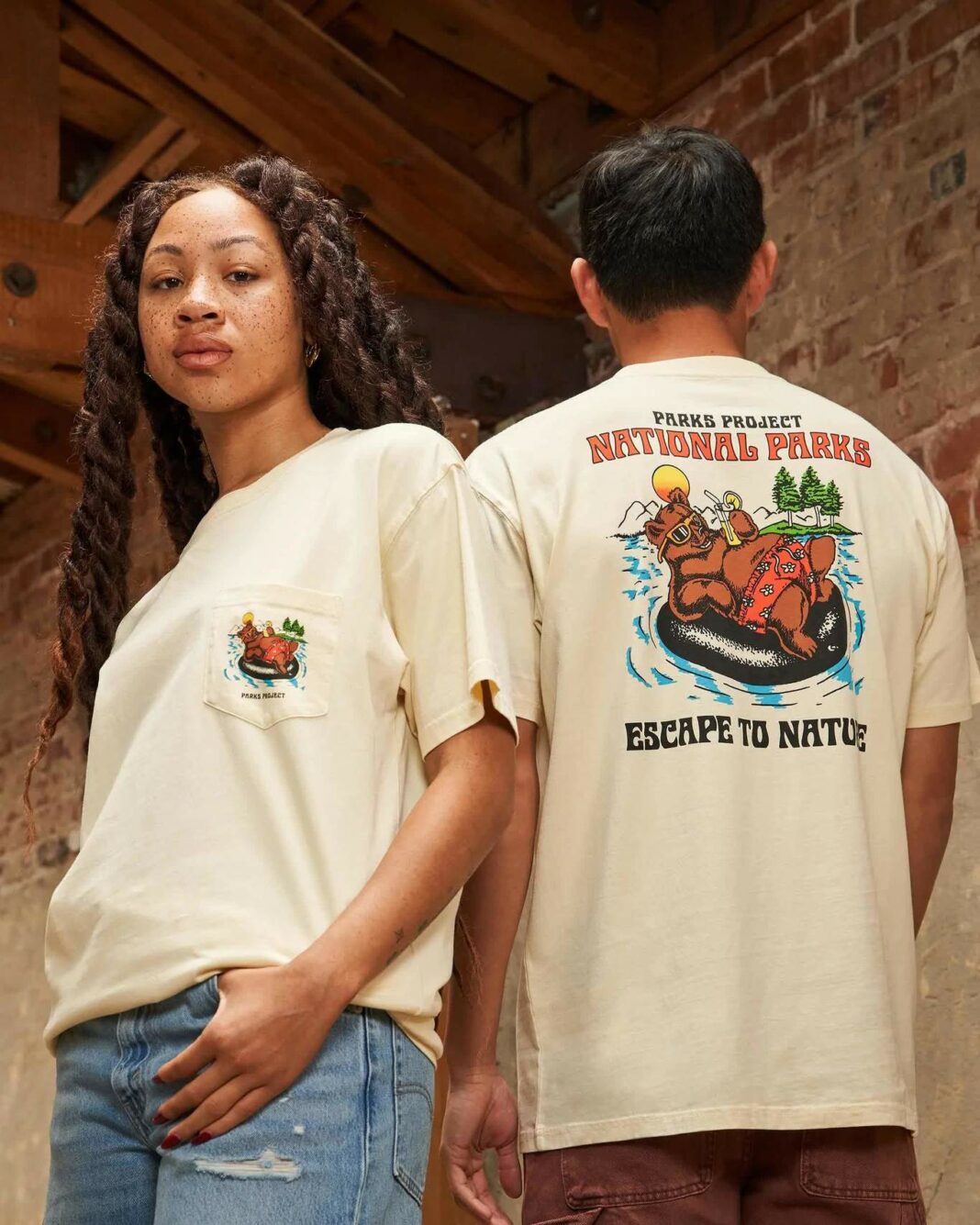 Two people in their 20s wear matching ivory tee-shirts that depict a bear in sunglasses floating on an inner tube in front of trees. The shirt reads “Parks Project / National Parks / Escape to Nature”